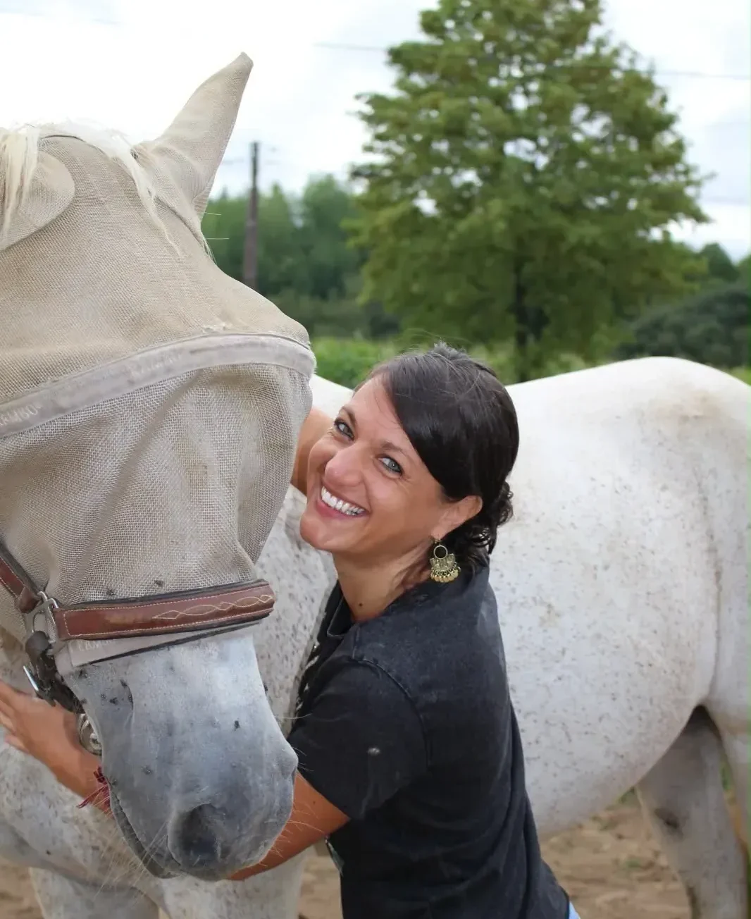 Praticienne entourant l’encolure d’un cheval gris et souriant vers l’objectif, le cheval portant un masque anti-mouches, tourne la tête vers elle.